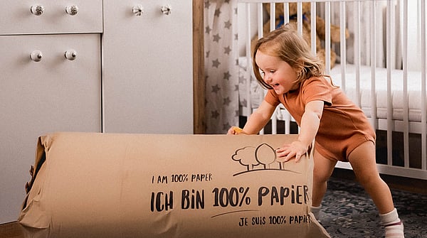 young girl in orange t-shirt and orange shorts leaning on a large piece of cardboard packaging and smiling with a crib behind her